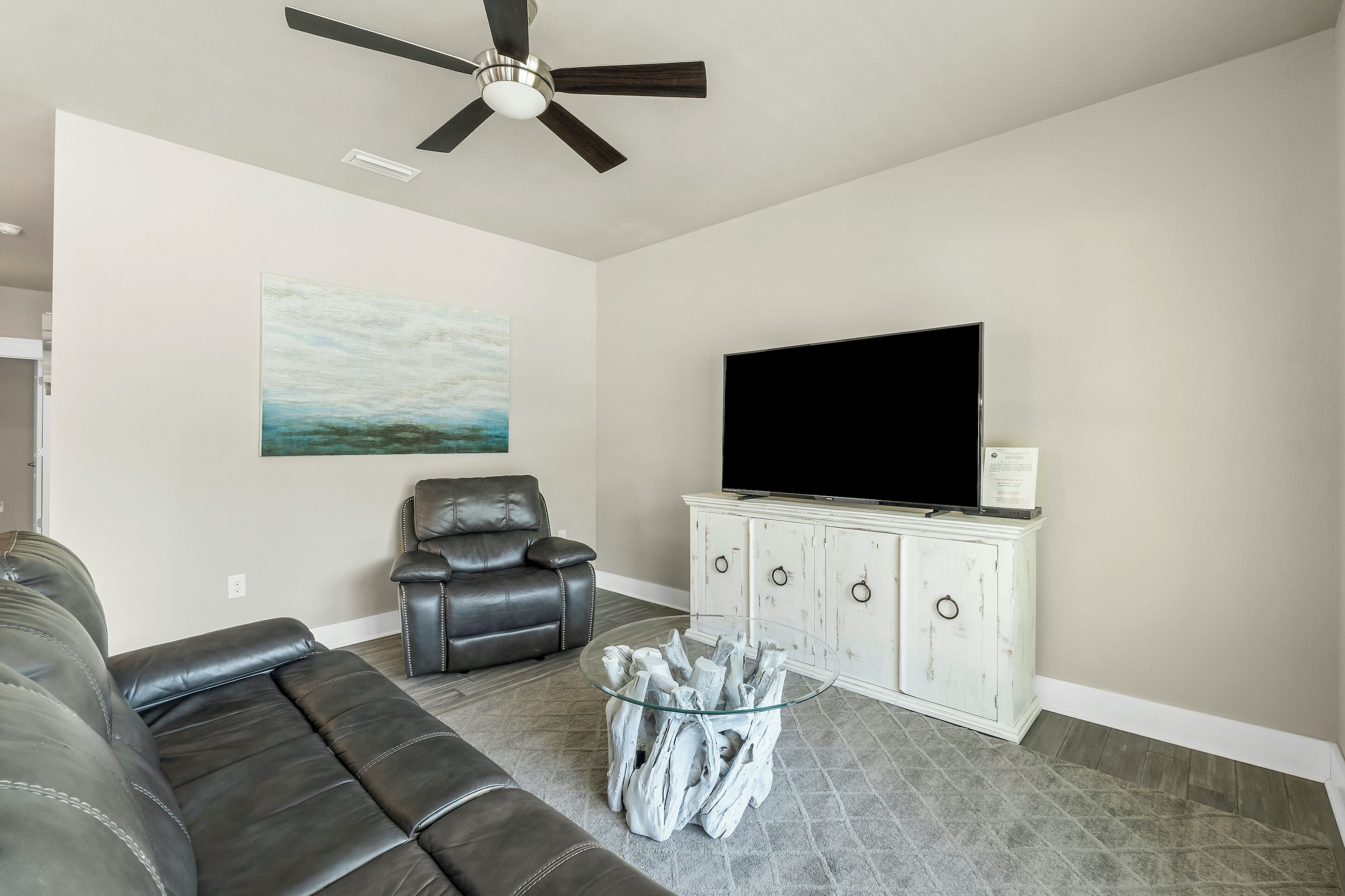 Una sala de estar con un sofá de cuero negro, una mesa de centro de vidrio, un mueble blanco con un televisor de pantalla plana y un ventilador de techo.
