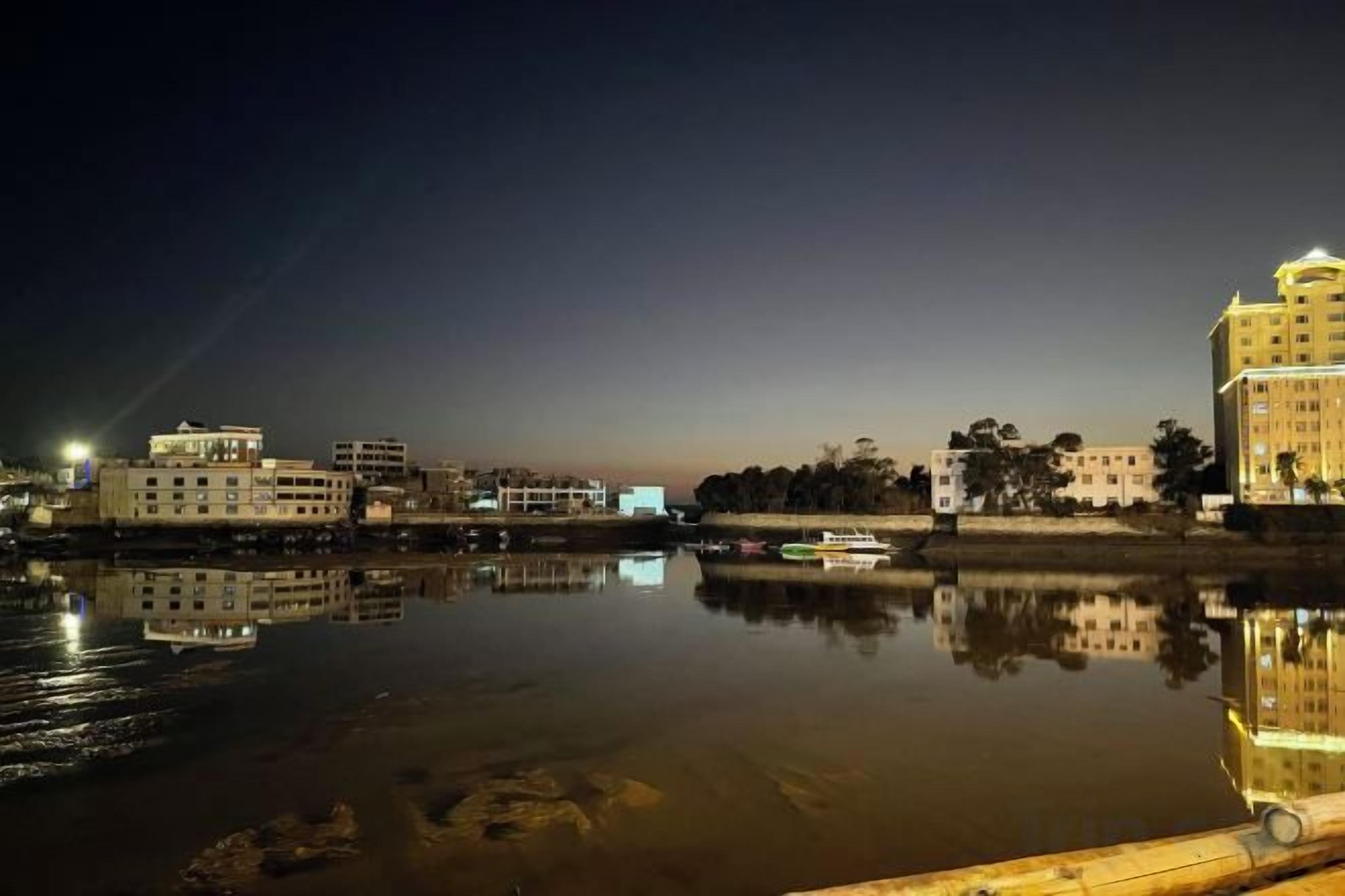 A nighttime cityscape with buildings reflected in calm water, a dock, and a boat.