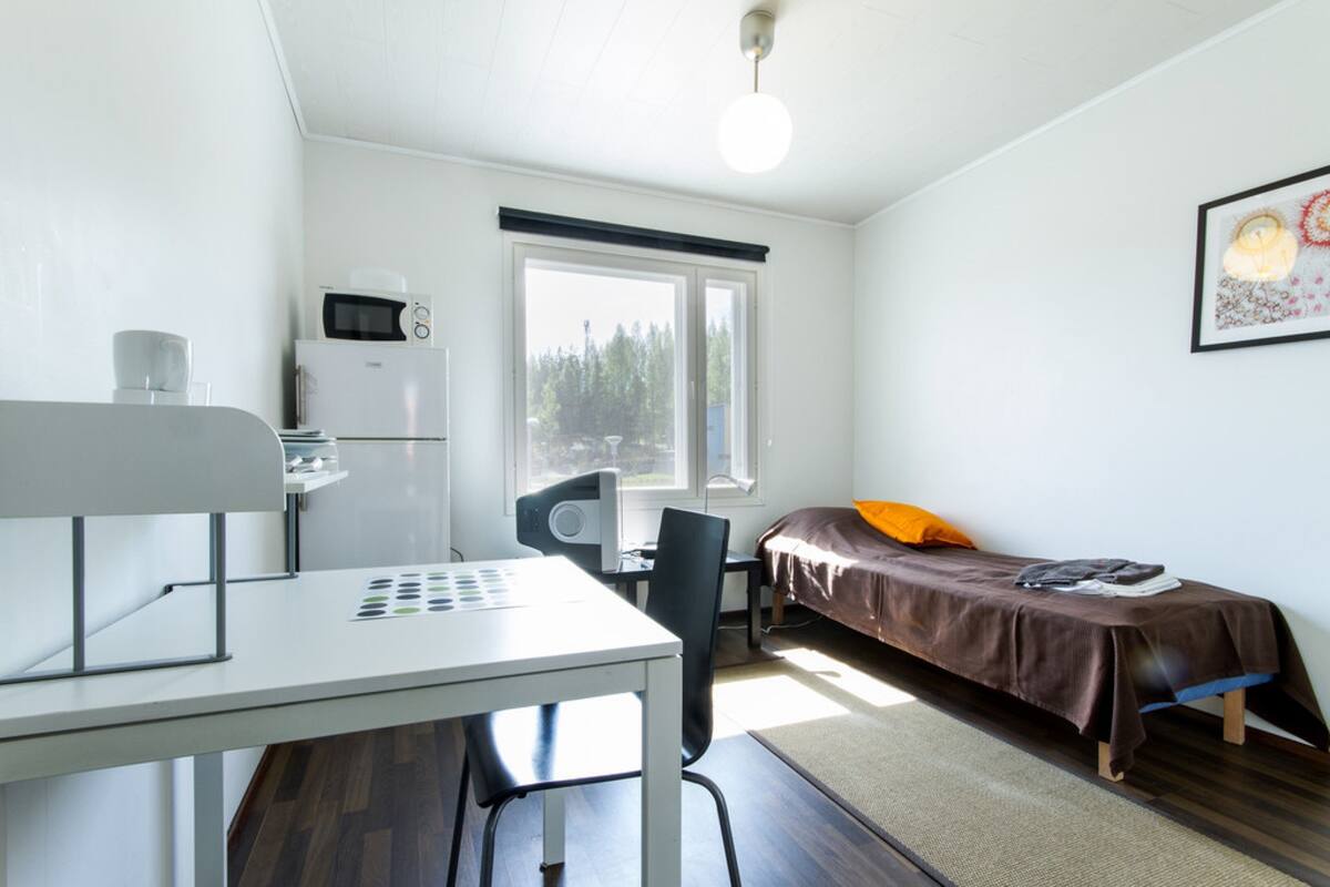 A small, well-lit room with a bed, a desk, a chair, a microwave, and a refrigerator.