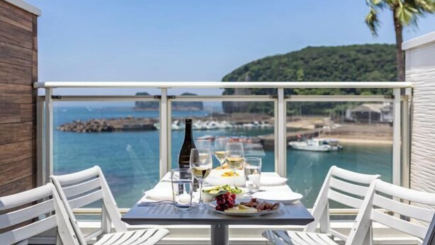 Un balcon avec une table dressée pour le dîner, donnant sur une zone côtière avec des bateaux et une colline boisée.