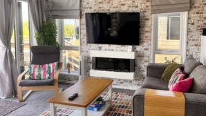 Een woonkamer met een bakstenen muur, een flatscreen televisie boven een open haard, een grijze bank met kleurrijke kussens, een houten salontafel en een veelkleurig tapijt.
