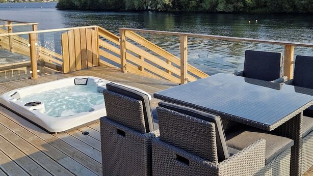 Een houten terras met een jacuzzi, rieten stoelen en een tafel aan een rustig waterlichaam, met bomen op de achtergrond.