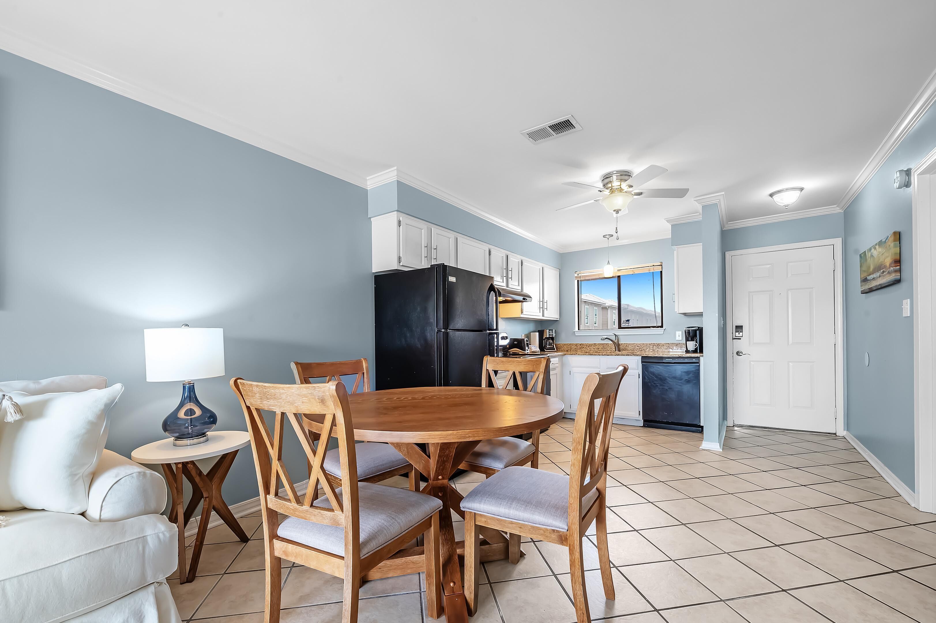 A compact kitchen with a dining area, featuring a round wooden table, chairs, a black refrigerator, and a ceiling fan.