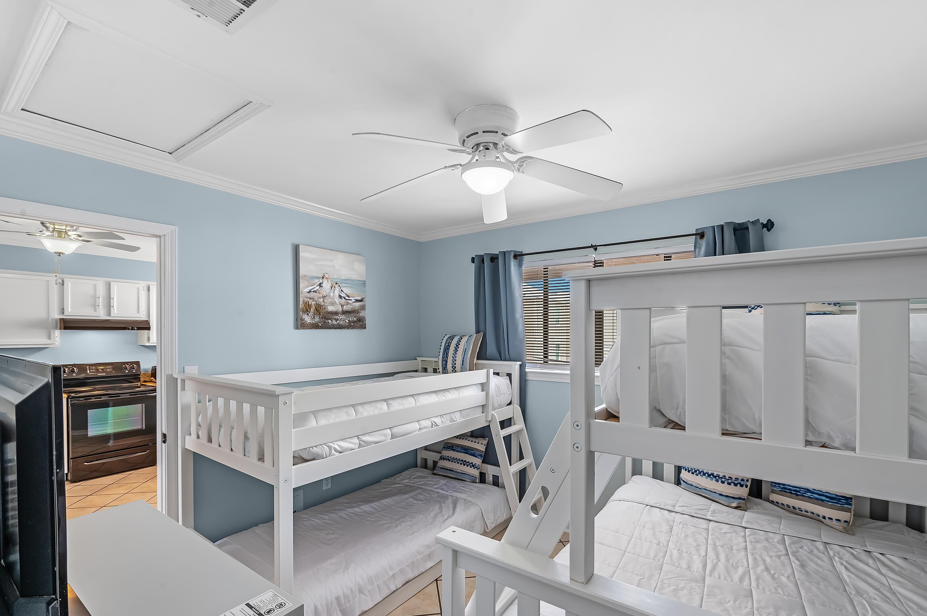 Un dormitorio con literas, ventilador de techo, zona de cocina y ventana con cortinas.