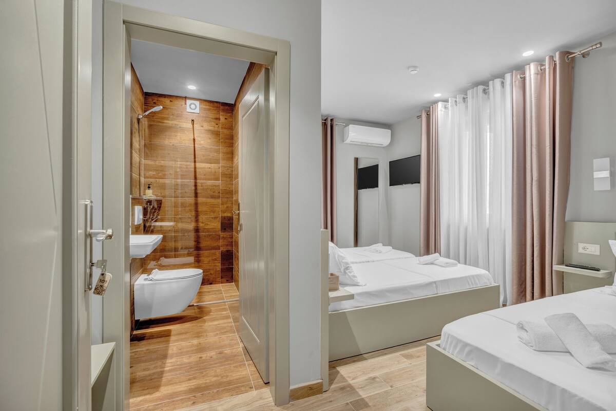 Une chambre d’hôtel moderne avec une douche en bois, un lavabo blanc, deux lits et une table de chevet avec une lampe.