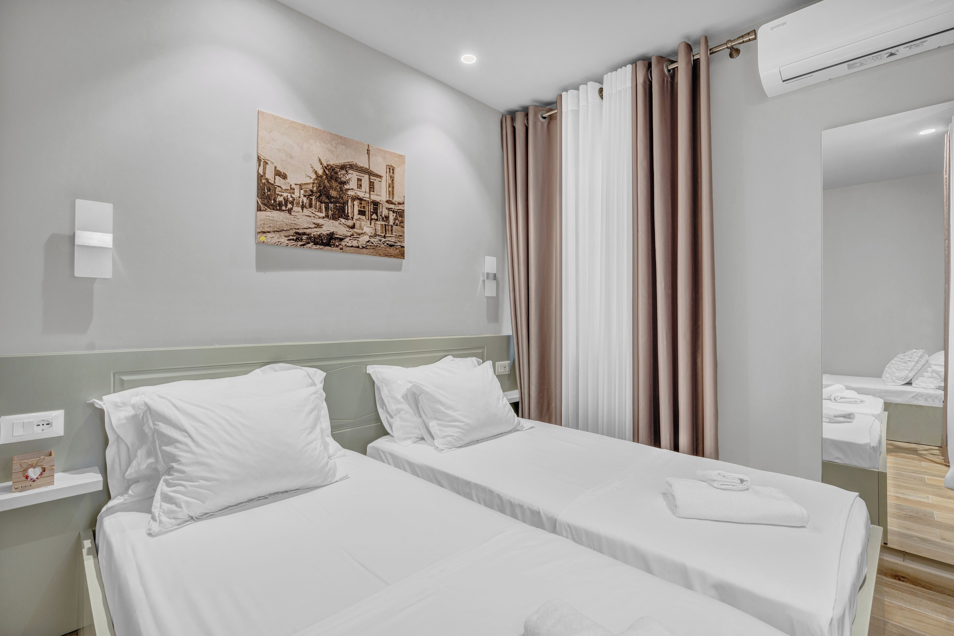 Una camera d'albergo con due letti singoli, un condizionatore d'aria a parete, uno specchio e un quadro raffigurante una scena di strada.