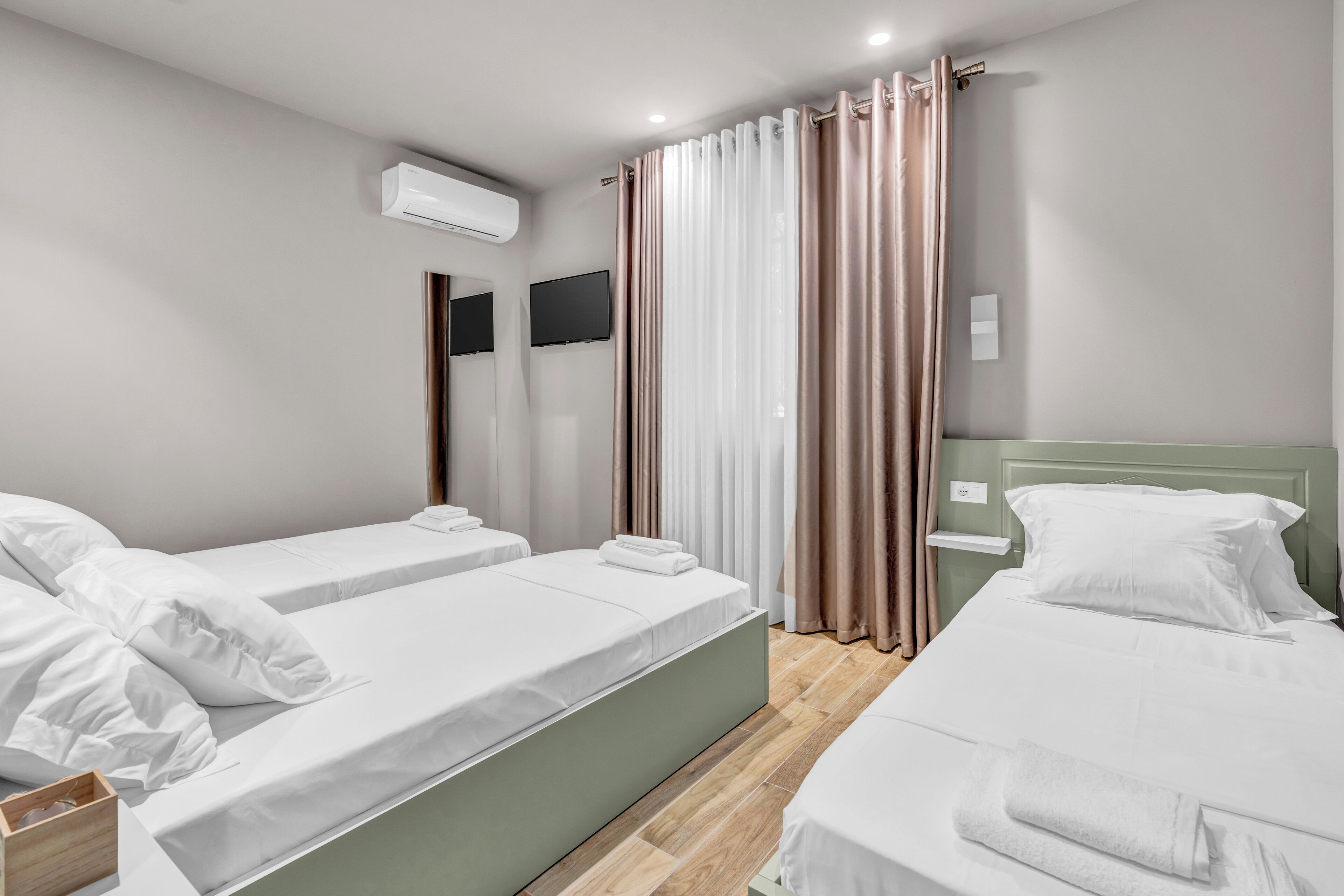 A hotel room with two beds, a flat-screen TV, an air conditioning unit, and a window with curtains.