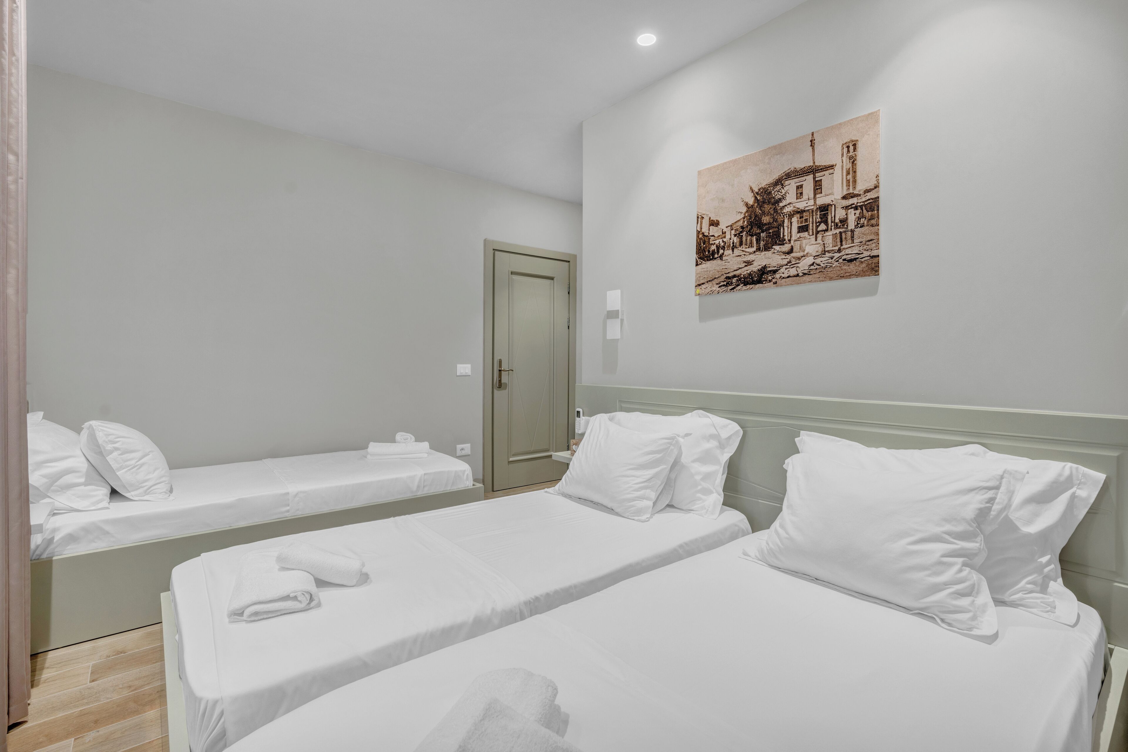 A hotel room with two single beds, a door, a wall-mounted painting, and a small bedside table.