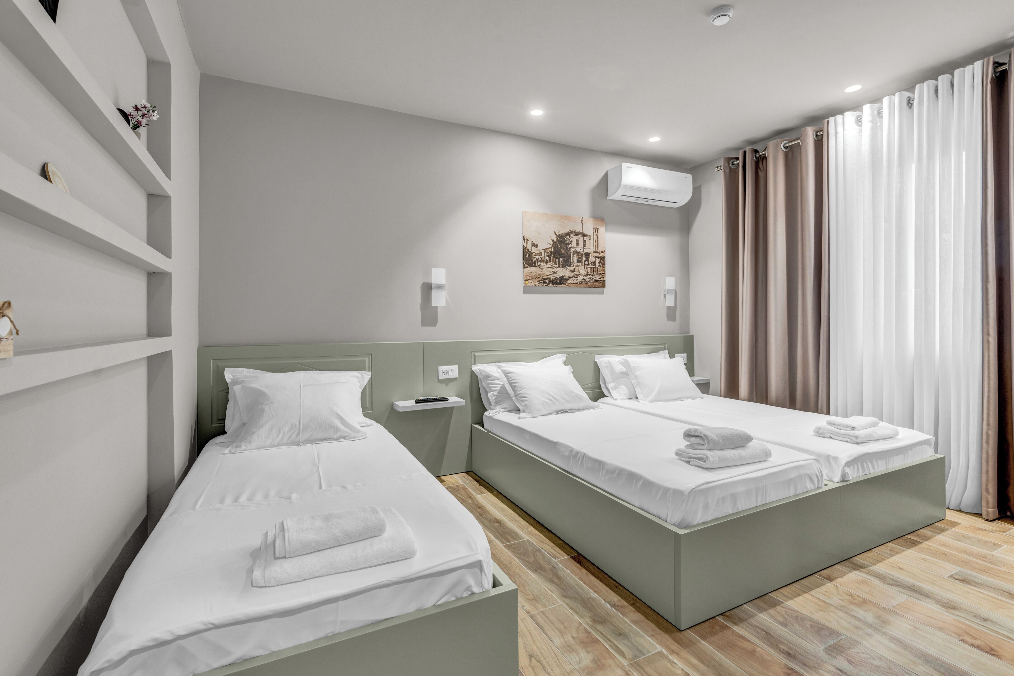 Habitación de hotel moderna con dos camas individuales, una mesita de noche pequeña, aire acondicionado de pared y un ventanal con cortinas.