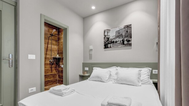 Un lit bien fait, avec du linge de lit et des serviettes blanches, une porte en bois, une petite table de chevet avec une lampe et une photographie en noir et blanc accrochée au mur.