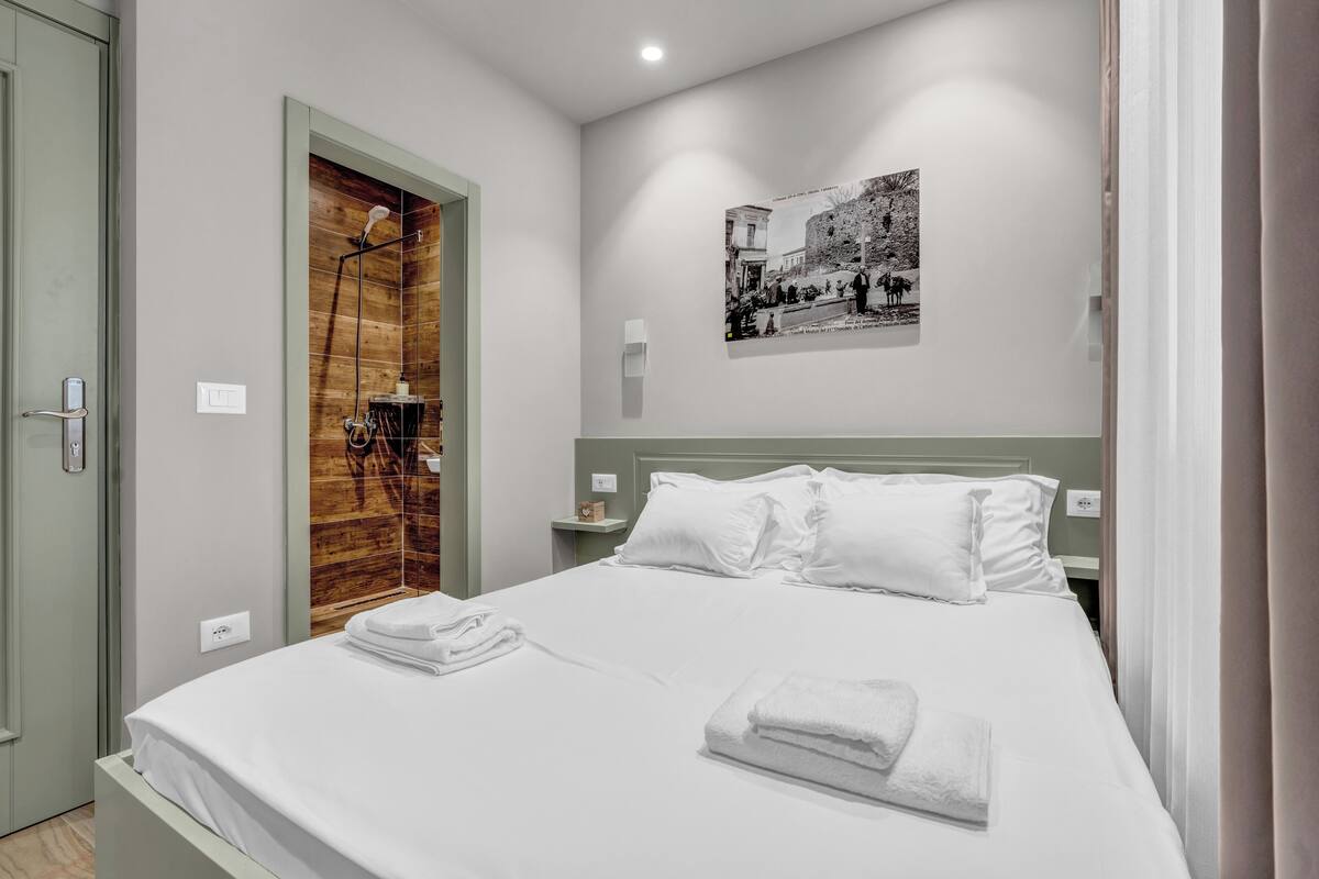 Un lit bien fait, avec du linge de lit et des serviettes blanches, une porte en bois, une petite table de chevet avec une lampe et une photographie en noir et blanc accrochée au mur.