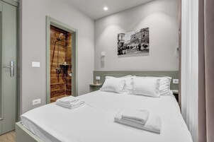 A neatly made bed with white linens and towels, a wooden door, a small nightstand with a lamp, and a black and white photo on the wall.