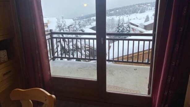 A room with a wooden floor, a wooden chair, a table, and a sliding glass door leading to a balcony with a view of a snowy landscape. - Apartment Les Saisies (Hauteluce)