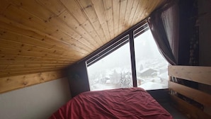A room with a wooden ceiling, a bed with a red and beige bedspread, and a window with a view of a snowy landscape. - Apartment Les Saisies (Hauteluce)