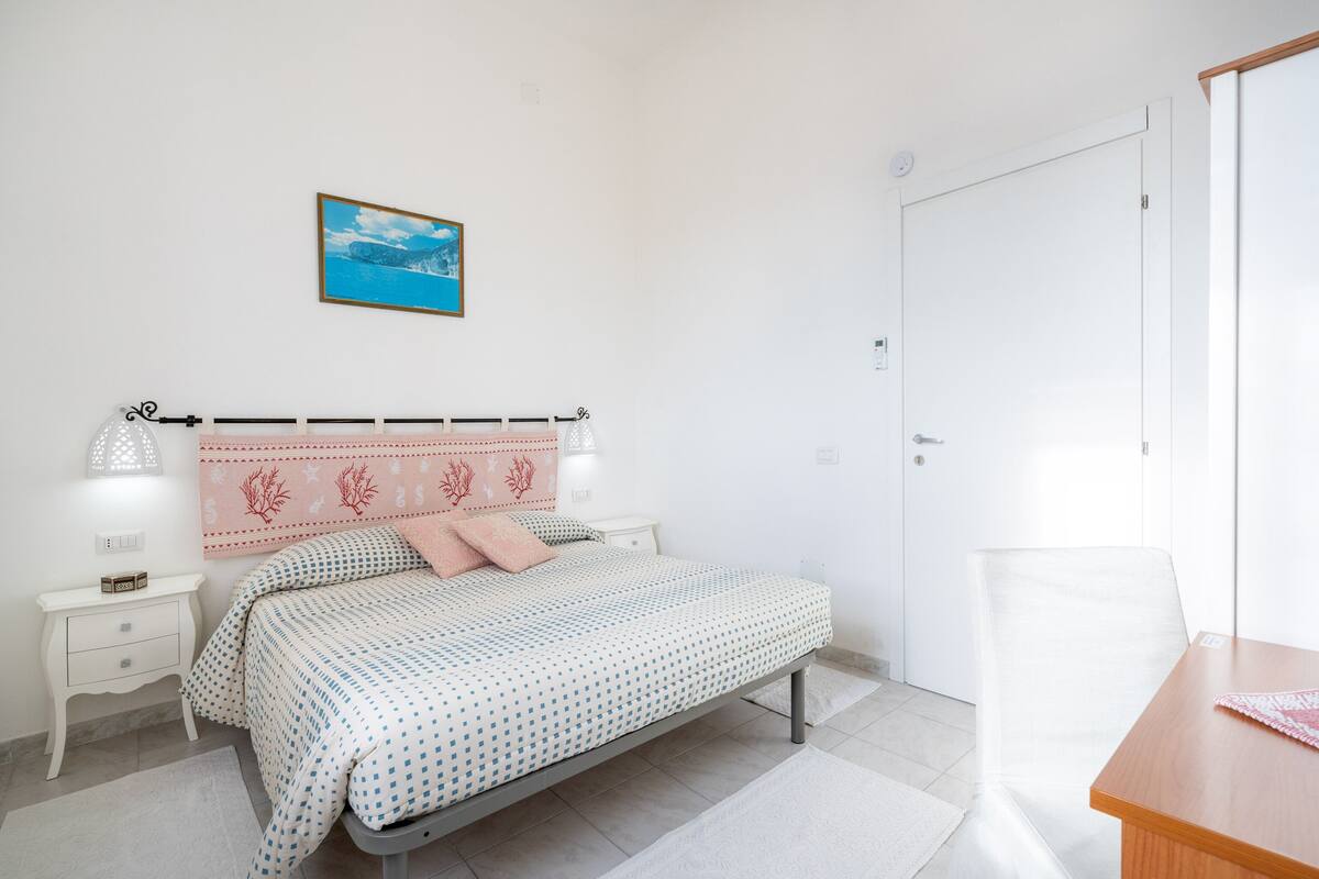 Ein Schlafzimmer mit Bett, Nachttischen, einem Sessel, einem Schreibtisch, einem Bild an der Wand und einer Tür.