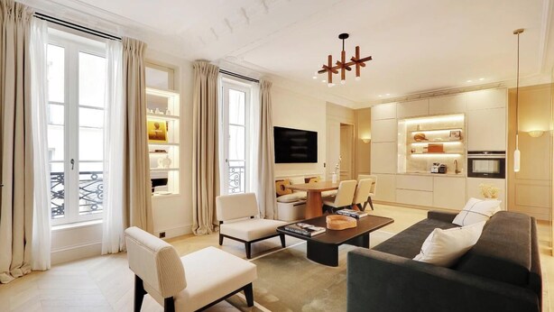 A modern living room with a sofa, armchair, and coffee table, a dining area with chairs, and a kitchen with built-in appliances. - EXQUISITE PARISIAN LUXURY: SOPHISTICATED RETREAT NEXT TO LOUVRE - RUE DE RIVOLI (Paris)