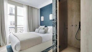 A bedroom with a bed, a nightstand, a window with curtains, and a view of the outside. - EXQUISITE PARISIAN LUXURY: SOPHISTICATED RETREAT NEXT TO LOUVRE - RUE DE RIVOLI (Paris)