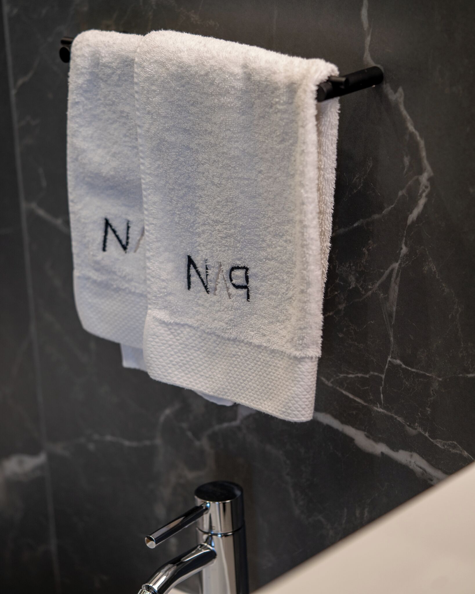 Een badkamer met een marmeren wand, een zwarte kraan en twee witte handdoeken met de letters 'N' en 'P' geborduurd erop.