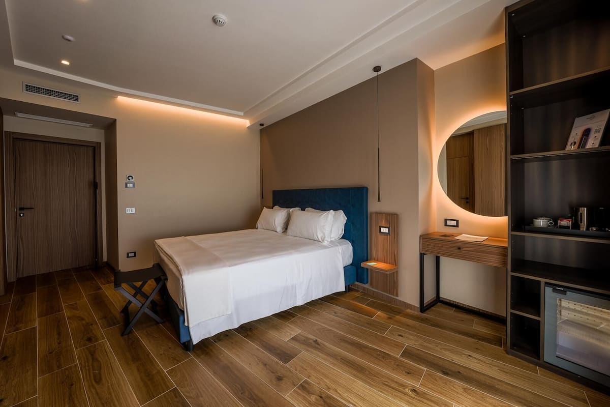 A modern hotel room with a large bed, a wooden wardrobe, a built-in desk, and a mirror.