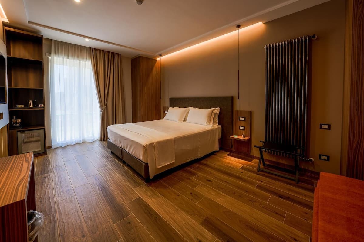 A modern hotel room with a large bed, wooden flooring, a built-in wardrobe, a desk, and a radiator.