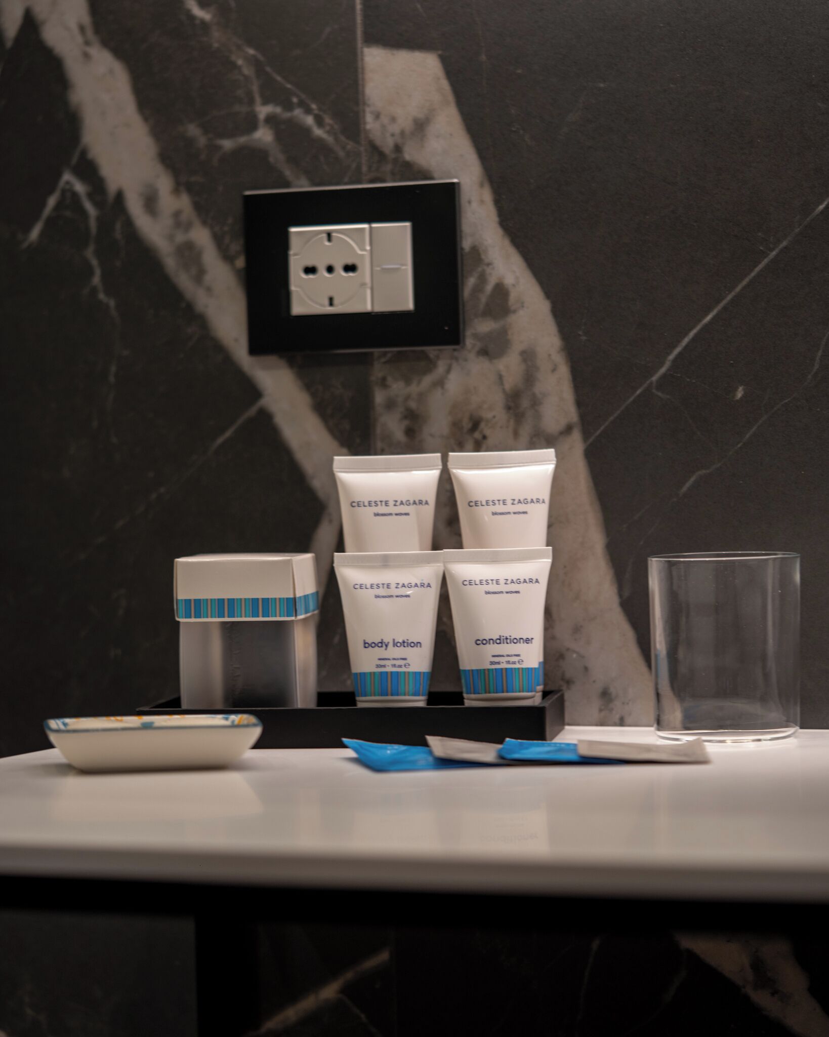 A bathroom counter with toiletries, a glass, and a marble wall.