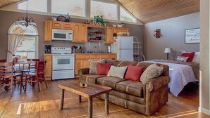 A spacious living area with a wooden ceiling, a kitchen with wooden cabinets, a dining table with chairs, and a cozy bed with a red pillow.