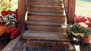 A wooden staircase leading to a porch with a metal railing, surrounded by potted plants and a doormat.