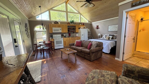 A cozy living room with a wooden ceiling, a dining area with a table and chairs, a kitchen with wooden cabinets and appliances, and a bedroom with a bed and a door leading outside.