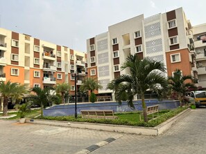 Un immeuble résidentiel doté de nombreux balcons, d’une cour pavée agrémentée de palmiers et d’un banc.
