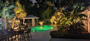 A pool area with a covered seating area, surrounded by lush tropical plants and trees.