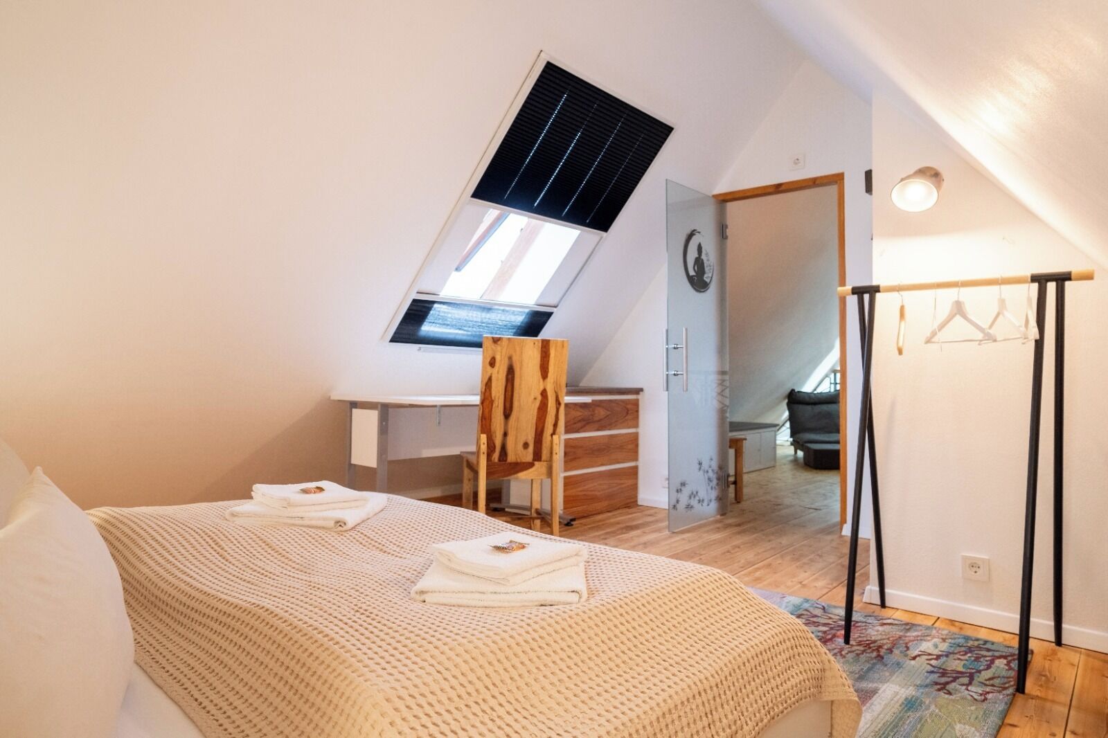 Ein Schlafzimmer mit schrägen Wänden, einem Oberlicht, einem Bett mit beiger Decke, einem Holzstuhl, einem Schreibtisch und einem Kleiderschrank.