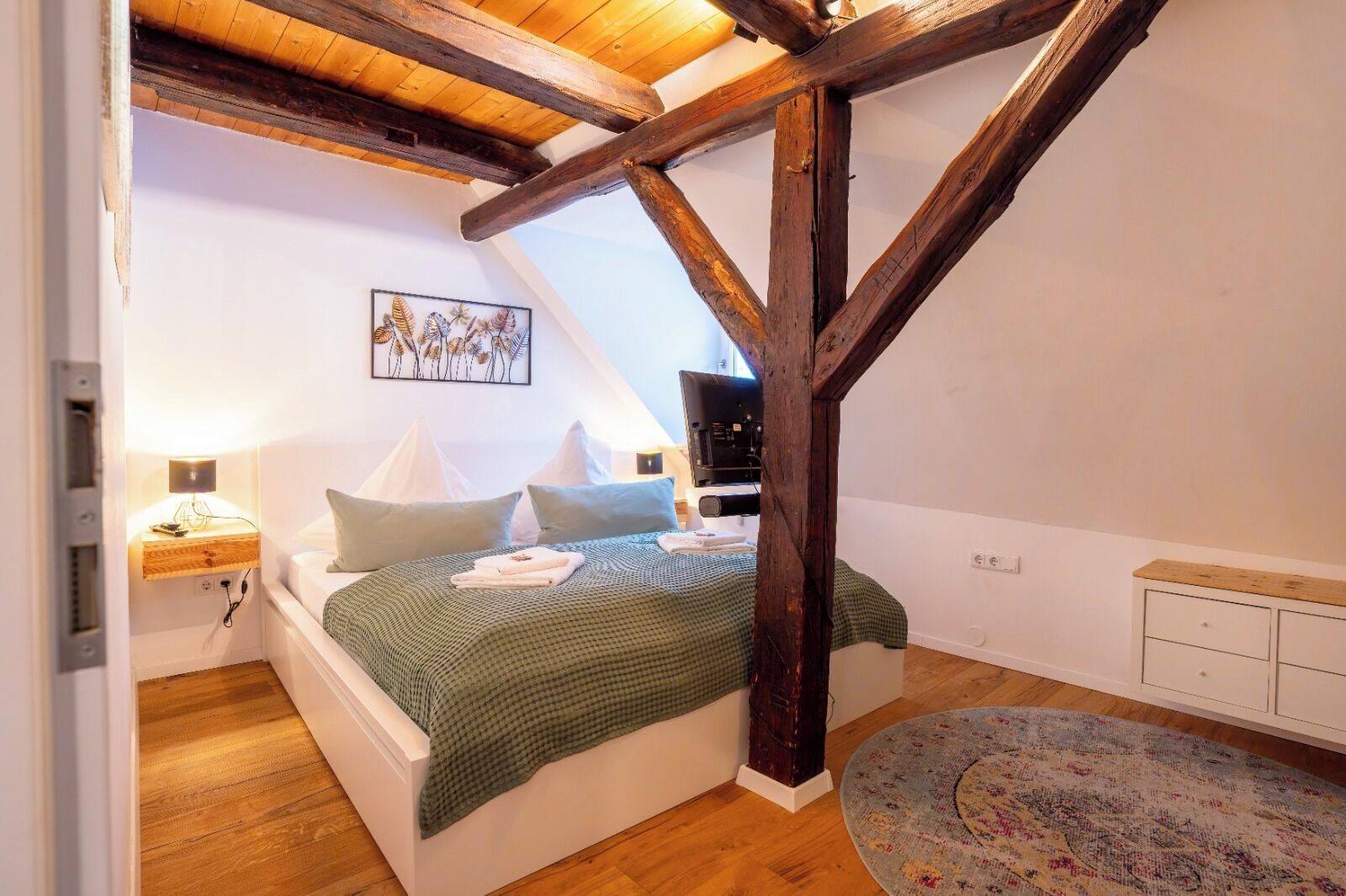 A bedroom with a sloped ceiling, wooden beams, a bed with pillows, a bedside table with a lamp, a television, and a patterned rug.