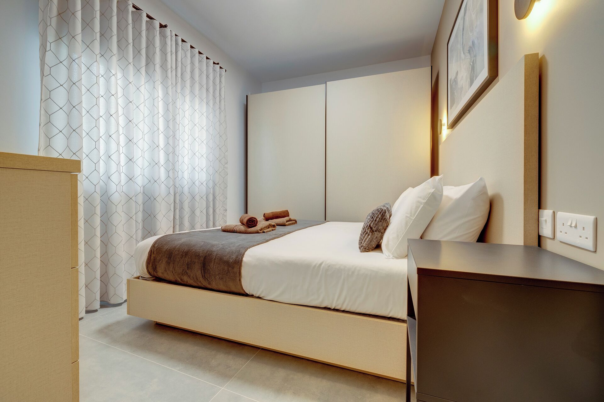 A modern hotel room with a bed, a bedside table, a wall-mounted light, and a curtain with a geometric pattern.