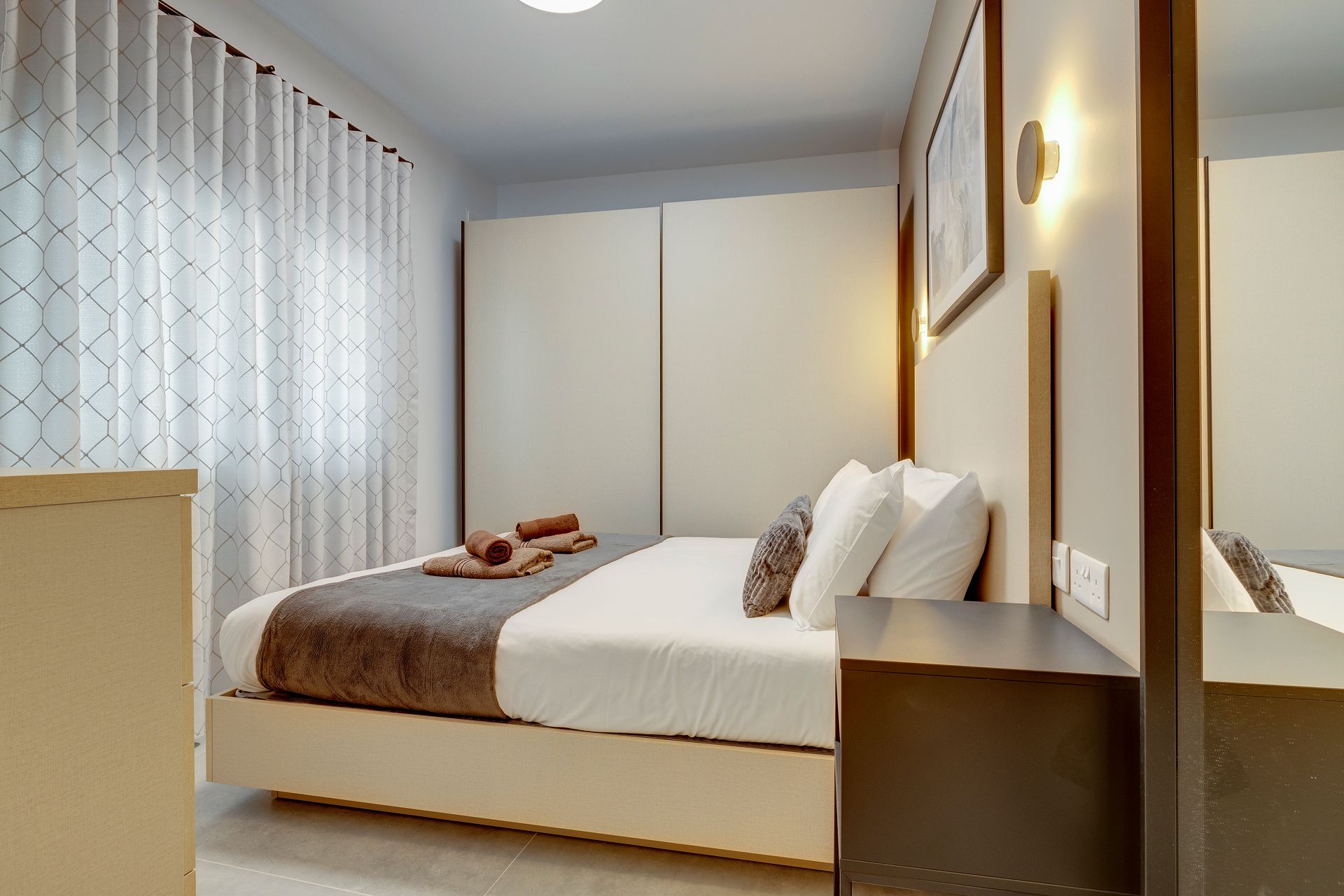 Une chambre à coucher moderne avec un lit, une table de chevet, un miroir et une fenêtre avec des rideaux.