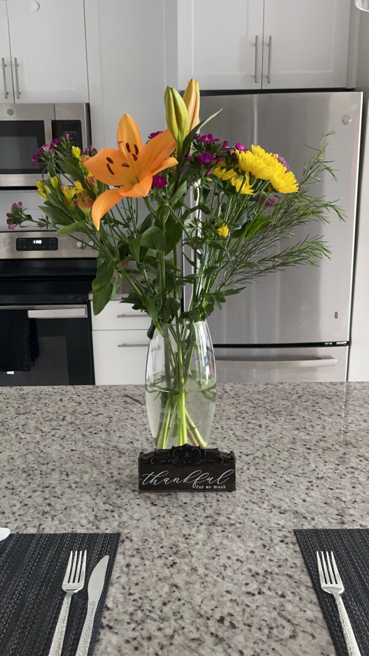 Un vase rempli de fleurs est posé sur le comptoir de la cuisine, et une petite pancarte à côté indique : « Je suis reconnaissant(e) pour tout. »
