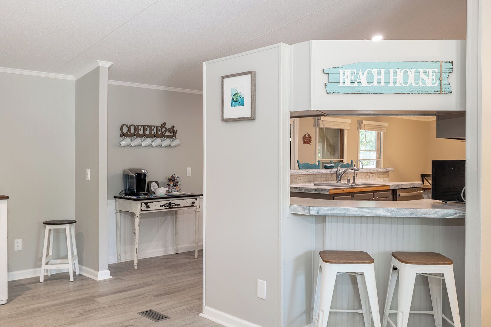 Une cuisine équipée d’un coin café, une pancarte indiquant « MAISON DE PLAGE », et un bar avec des tabourets.