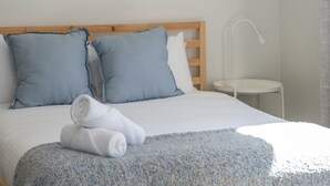Ein ordentlich bezogenes Bett mit blauen Kissen, einer weißen Decke und aufgerollten Handtüchern auf einem strukturierten blau-weißen Teppich.