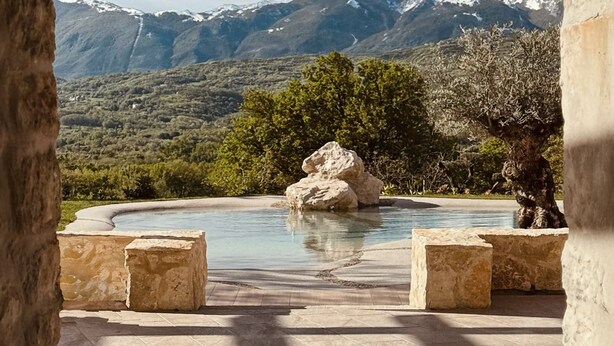 Une piscine agrémentée d’un élément central en roche, entourée de bancs en pierre, avec un paysage montagneux en arrière-plan.