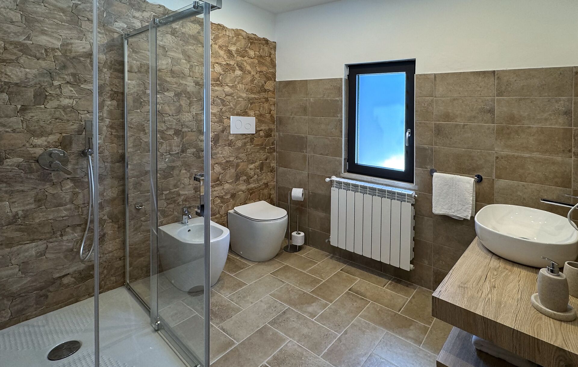 Un baño moderno con ducha envuelta en vidrio, dos bidés, un lavamanos grande y una ventana sobre el espejo.