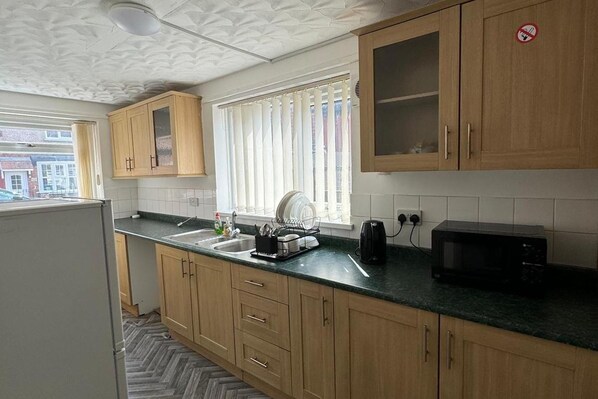 A kitchen with wooden cabinets, a sink, a microwave, and a window with blinds. - Hartlepool - 2 Bedroom - Near Seaside - Sleeps 5 (Hartlepool)
