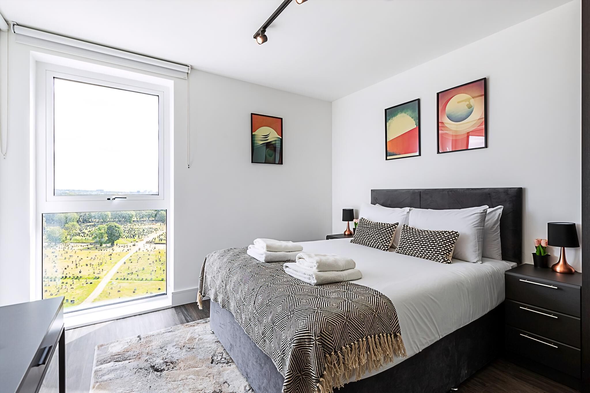 A bedroom with a large window, a bed with patterned bedding, two wall art pieces, and a nightstand with a lamp.