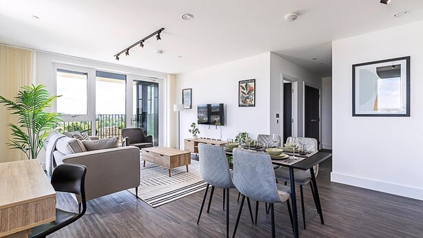 Een moderne woonkamer met een bank, eettafel en stoelen. Er hangt een televisie aan de muur en er is een raam met gordijnen.