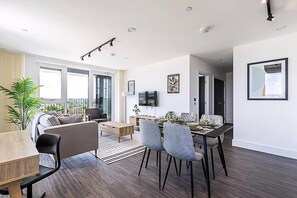 A modern living room with a sofa, dining table, and chairs. There is a TV on the wall and a window with curtains. - Sleek and Stylish 9th Floor Apartment (London)