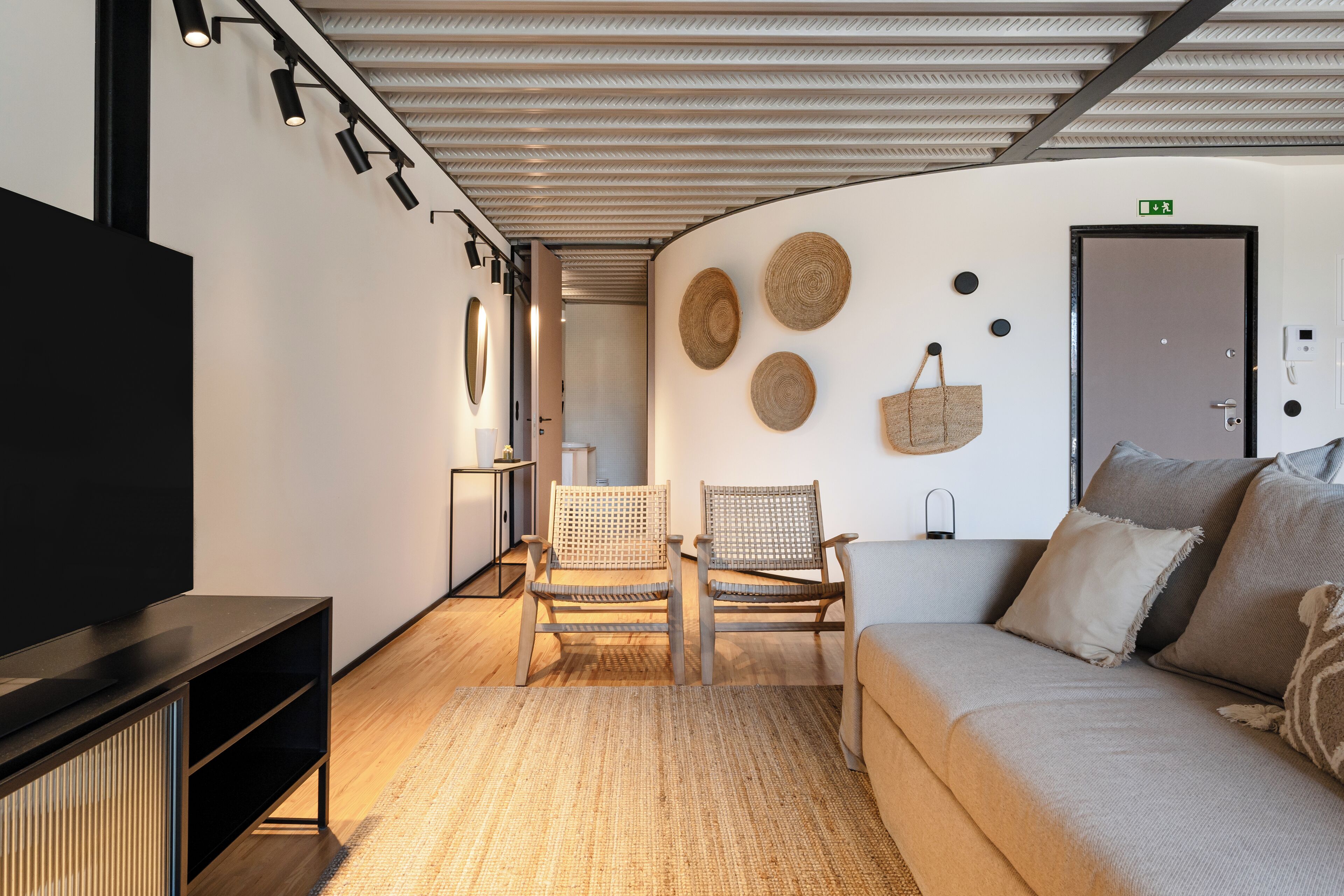 A modern hotel room with a flat-screen TV, a sofa, two wicker chairs, a woven rug, and a wall with woven wall art.