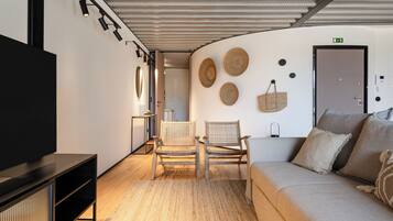 A modern hotel room with a flat-screen TV, a sofa, two wicker chairs, a woven rug, and a wall with woven wall art.