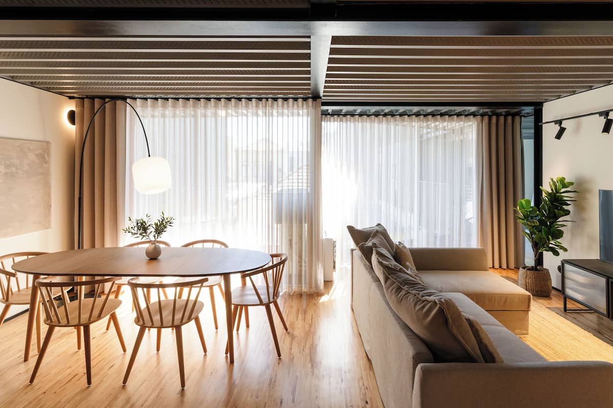 Un salon moderne avec une table à manger ronde en bois, un canapé beige et une grande fenêtre ornée de rideaux légers.