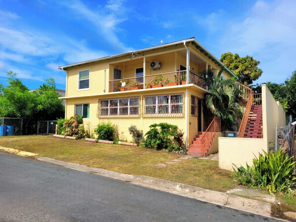 A two-story yellow building with a balcony, a staircase, and a small garden area.