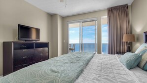 Une chambre d’hôtel avec un lit, un téléviseur posé sur une commode en bois, un balcon avec des rideaux et une vue sur la mer.