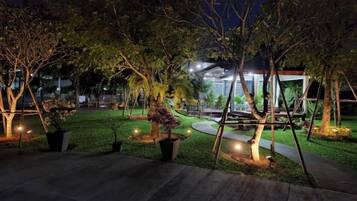 A nighttime outdoor area with trees, potted plants, and a pathway leading to a building.