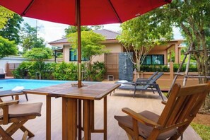 A poolside area with a wooden table and chairs, a red umbrella, and a view of a house with a tiled roof. - Baan Nern Khao Resort Pattaya (Pattaya)